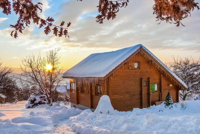 Snow-covered wooden alpine chalet at sunrise, with icicles hanging from the roof and frosted oak branches framing the scene