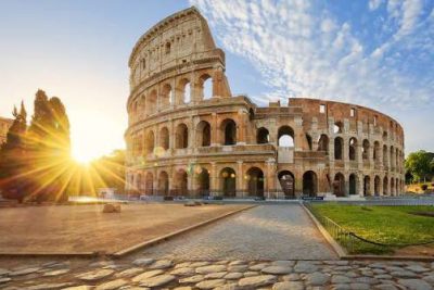 The Colosseum in Rome at sunrise, with golden light breaking through its ancient arches and an empty cobblestone street in the foreground